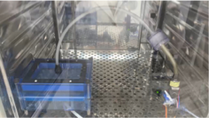 A blue rectangular object connected to hoses inside a metal chamber with perforated grid floor, suggesting a scientific or industrial experiment.